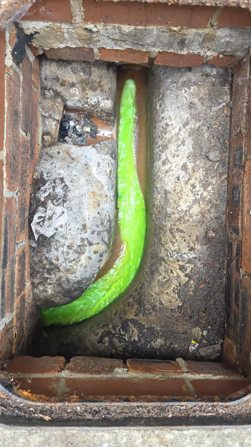 View down into a brick drainage inspection chamber showing bright green tracing dye flowing through the channel.